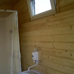 Wnętrze sanitariatu w drewnianej konstrukcji z uchylnym oknem, umywalką i dozownikiem mydła na ścianie z desek.